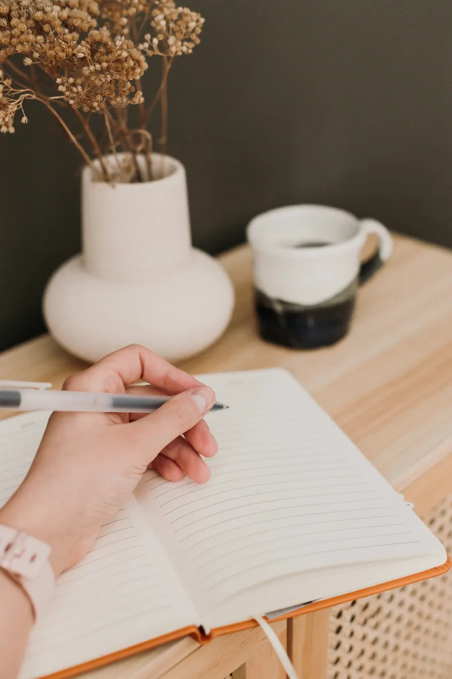 A hand writing in an open journal beside dried flowers and a ceramic mug
