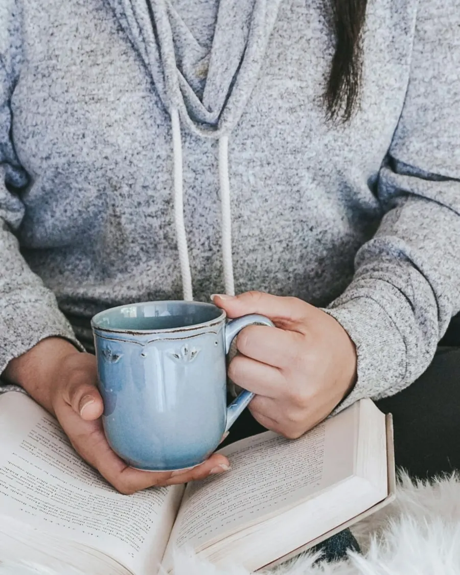 Someone in a soft grey jumper holding a blue mug over an open book
